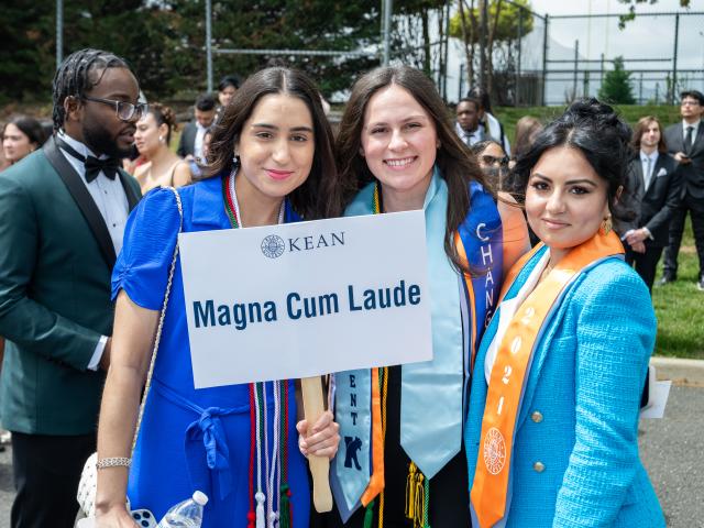 Honors graduates smile and hold up a Magna Cum Laude sign, as they wait for the convocation to begin