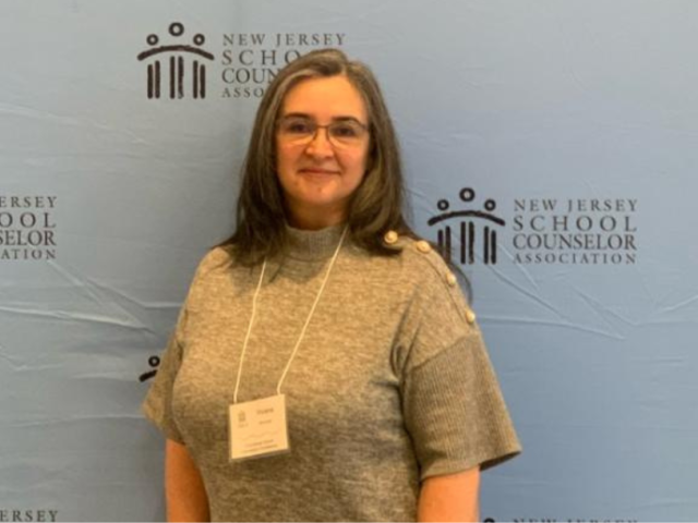 Woman with brown hair, glasses, a tan shirt and a conference name badge, in front of a light blue background