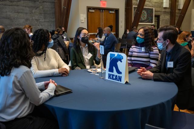 Five people of different ethnicities and races seated at a table with a Kean logo as the center piece, dressed in suits and talking to each other.