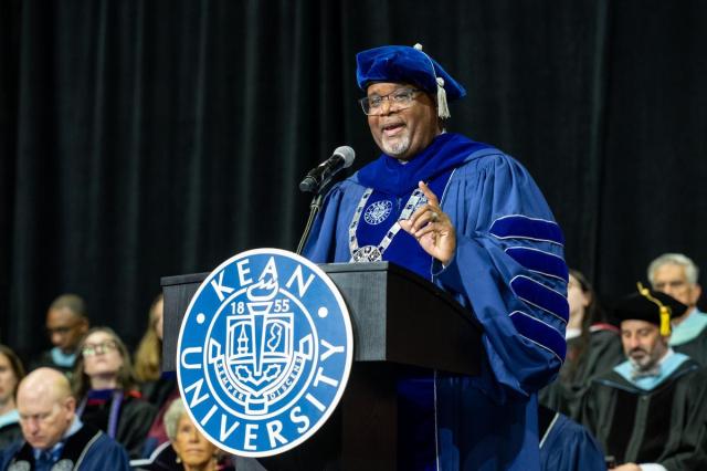 Black male wearing a blue graduate cap and gown, speaking at a Kean podium.