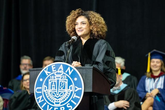 Young Hispanic female with short curly hair, black graduation gown, at a podium.