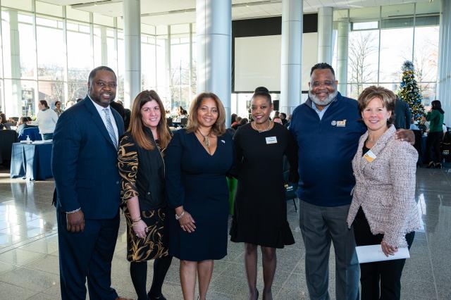  (L-R) A Black male with black hair and facial hair wearing a navy suit, a White female with long, brown hair wearing a long-sleeved brown and gold dress, a Black female with light brown hair, wearing a long-sleeved navy blue dress, a Black female with dark brown hair in a bun, wearing a long-sleeved black dress, President Lamont Repollet, a Black male with curly short black hair, wearing gray pants a navy blue top, and a white female with short brown/blonde hair, wearing a white blazer with black pants