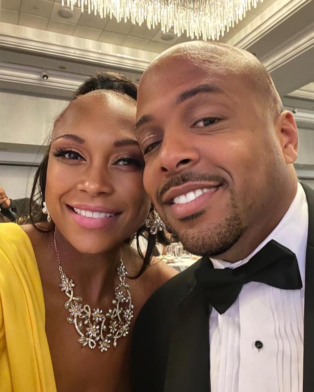 A Black woman wearing a yellow dress and a Black man in a tuxedo smile.