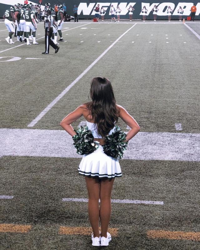 Megan Braine, in her Jets Flight Crew uniform, has her back to the camera and stands along the sidelines at at Jets game.