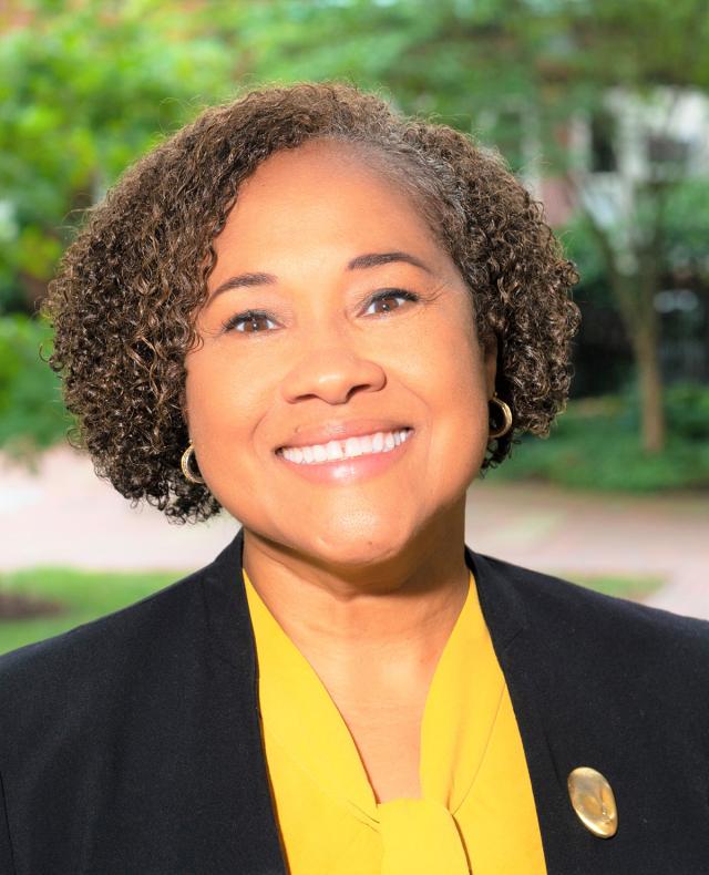 Rajade Berry-James, in a yellow shirt and black jacket, smiles broadly in a headshot.