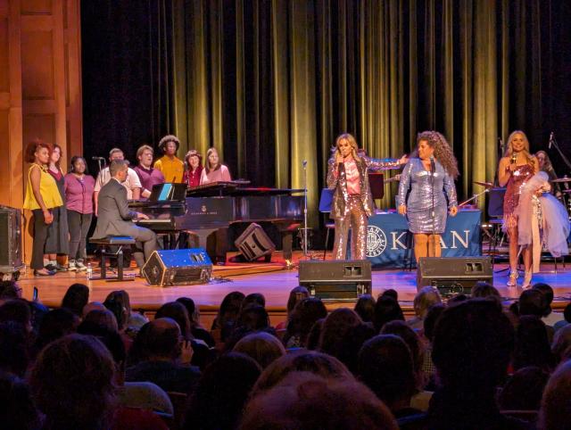 A diverse group of students on a stage to the left, to the right three adult women in sparkly outfits