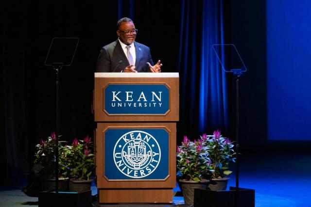 President Repollet gestures behind at podium at Wilkins Theatre