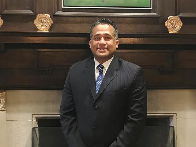 Alex Guzman, wearing a blue suit, stands in front of a fireplace.