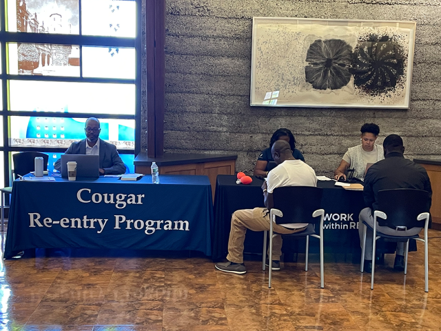 Two men, their backs to the camera, sit at a desk with the sign, "Cougar Re-entry Program."