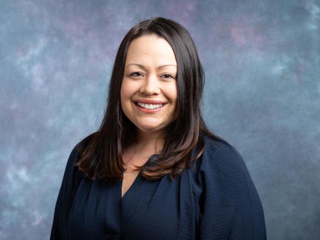 Neva Lozada Headshot - a Latino woman will long brown hair in a navy blue blouse smiles