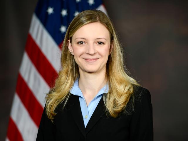 Ensela Mema, a white woman with long blond hair wears a black jacket over blue shirt and stands in front of an American flag.