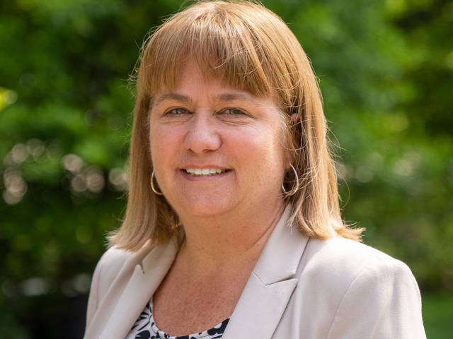 Sue Porterfield, a white woman with short light brown hair, in a beige jacket and floral shirt, standing outdoors in front of greenery.