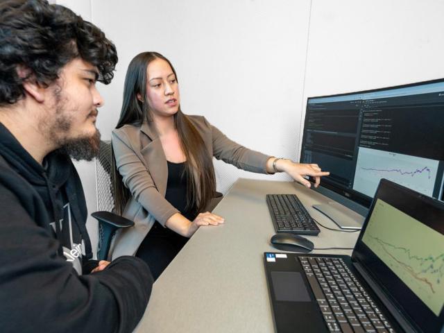 A young man and women sit before a computer at a desk. The woman is pointing to the screen.