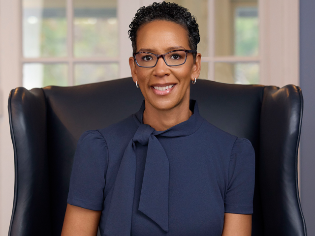 Darlene Repollet, with short black hair and glasses and wearing a dark blue suit, smiles as she sits in an upholstered chair.