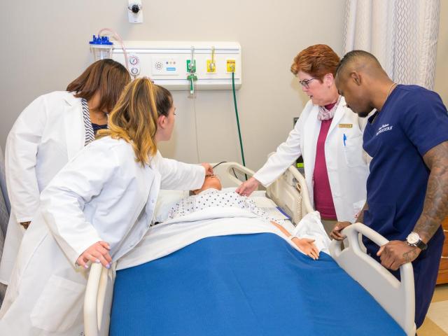 Three women and one man in nursing uniforms around a bed with a mannequin patient.