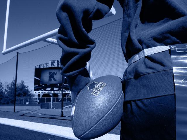 Upclose photo of a flag football player holding a Kean University football on a field; blue.