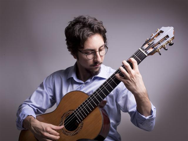 A young man with a light shirt playing the guitar within a gray background