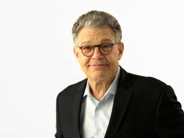 Portrait of Al Franken, former U.S. Senator and comedian, wearing glasses, a light blue collared shirt, and a black blazer. He is smiling softly