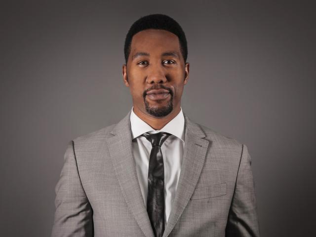 Portrait of Ndaba Mandela, grandson of Nelson Mandela, wearing a light grey suit, white shirt, and a dark tie. He has a short beard and mustache