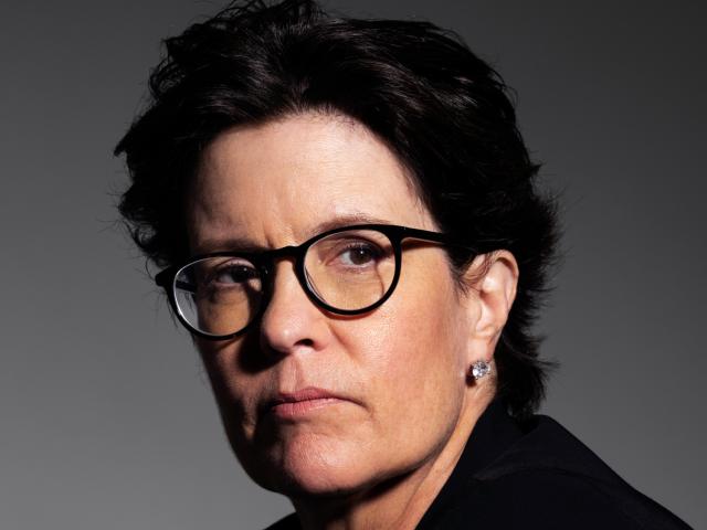 Portrait of Kara Swisher, prominent tech journalist and podcast host, wearing glasses with a dark frame, a black blazer, and a white top. She has short, dark hair and is looking slightly to the side with a serious expression. 