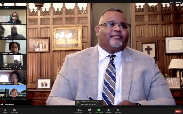 A screen shot of a Zoom event. Dr. Lamont Repollet speaks to First-Generation Students via Zoom. He is a Black male wearing a white shirt, gray suit jacket and striped tie, sitting in front of a wood-panel wall. Six people appear in Zoom boxes to the left of the frame.