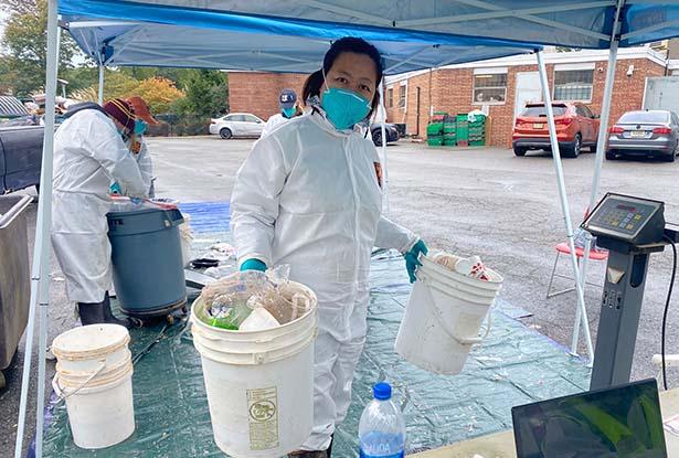 Kean professor Dongyan Mu in protective clothing hold two pails of sorted trash under a blue tent.