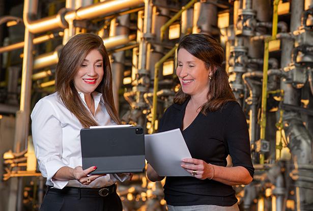 Two businesswomen standing before a wall of machinery and pipes; one holds a tablet, the other holds papers.