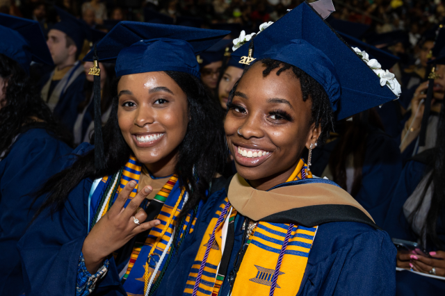 Two Black women, in Kean blue graduation caps and gowns, smile as they sit next to each other at Commencement.