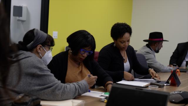 Three women and a man sit along a long desk, working together on computers.