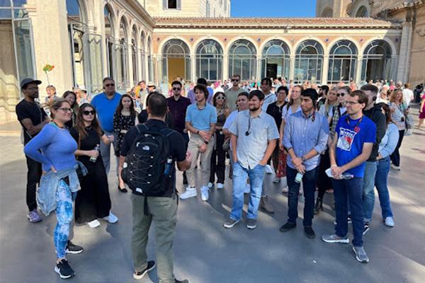 A group of students stand in a Roman plaza, facing their instructor.