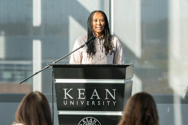  Chamique Holdsclaw, a Black female with long, black dreads standing behind a black podium that says Kean University on it in white. There is a black microphone in front of her as well. Chamique is wearing a cream-colored buttoned-up shirt, and is looking into the crowd. 