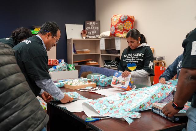 Three diverse people wrap Christmas gifts around a long table