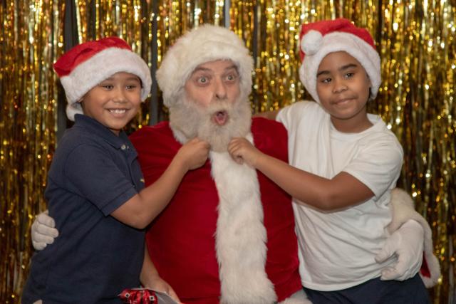 Two little boys wearing Santa hats sit next to Santa and pull on his beard.
