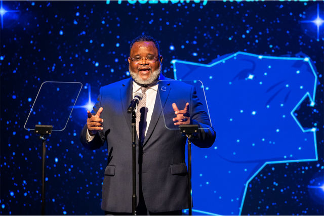 President Repollet stands before a screen of stars in the night sky, with a constellation of a cougar.