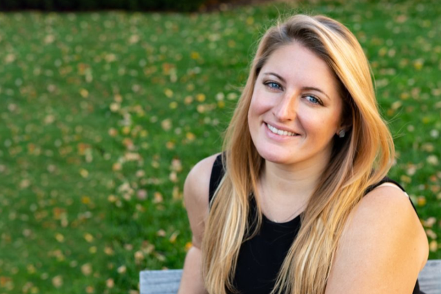 Kelly Bachovchin, a young blond woman, sits on a bench in a grassy area.