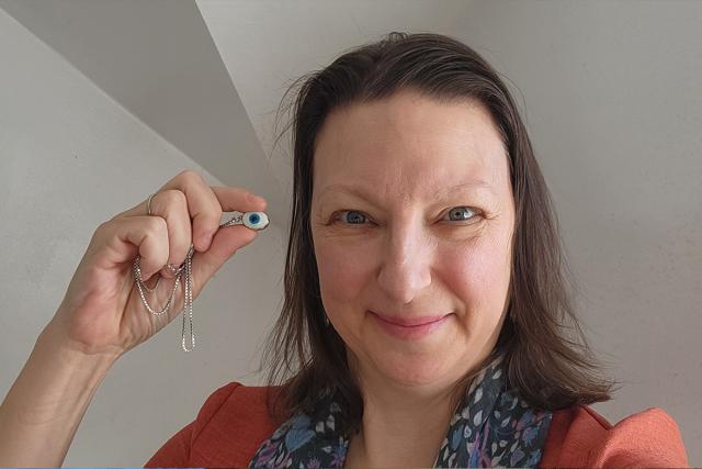 Smiling white female with brown hair, orange shirt and grey scarf holding up an evil eye amulet