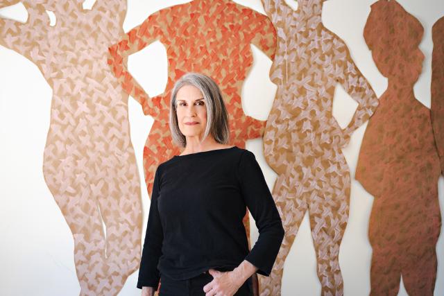 Woman with short gray hair wearing a black long-sleeved shirt standing in front of a mural painted with neutral-colored silhouettes of people