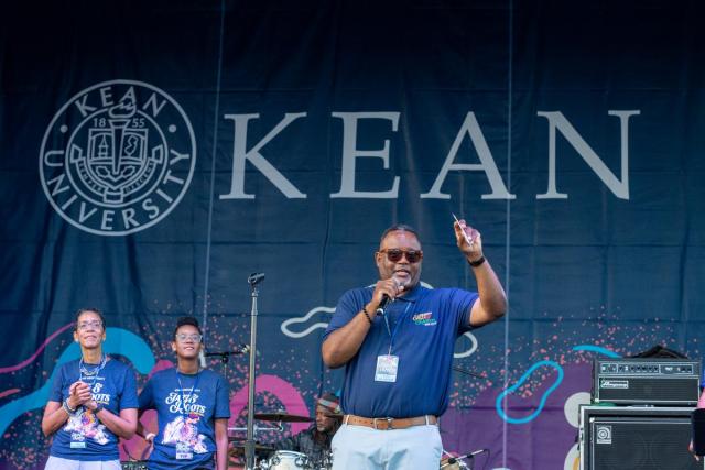 Kean President Repollet gestures as he speaks on stage; his wife and daughter are behind him.