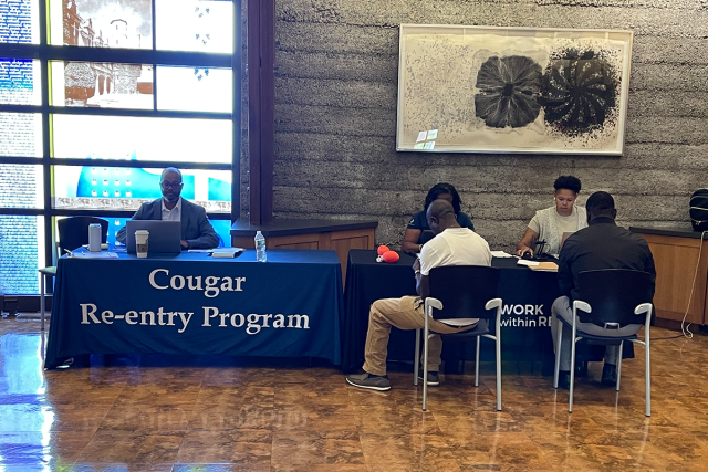 Two men, their backs to the camera, sit at a desk with the sign, "Cougar Re-entry Program."