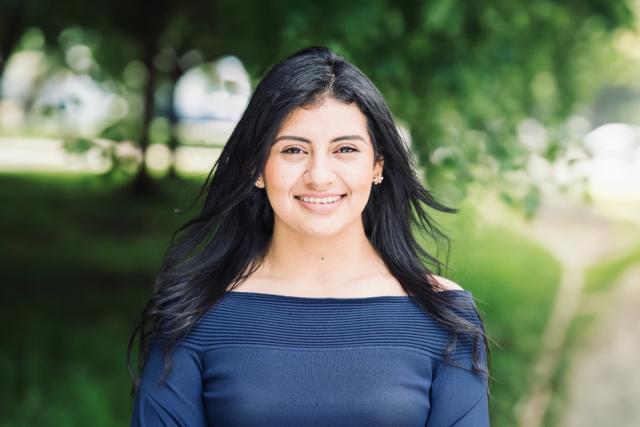 Diana Calle, with long dark hair and wearing a blue shirt, poses outdoors with trees behind  her.