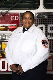 Rev. Pamela Holmes wearing white dress shirt, black pants and standing in front of a fire truck.