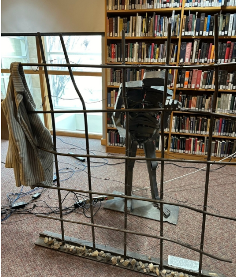 Holocaust memorial sculpture of figure behind prison fence. At the base of the sculpture are rocks symbolizing those that are commemorating Holocaust victims. On the fence is a concentration camp uniform shirt. Behind the sculpture are books at the Holocaust Resource Center.