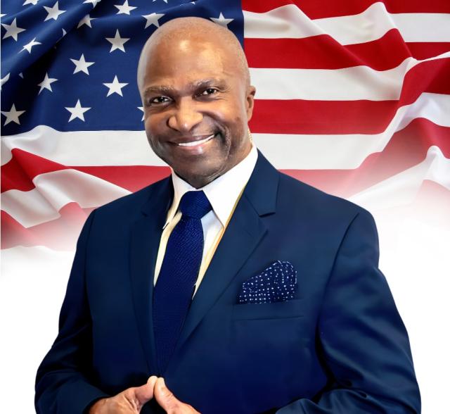 Mayor Adrian Mapp wearing a navy blue suit, smiling in front of an America flag background.