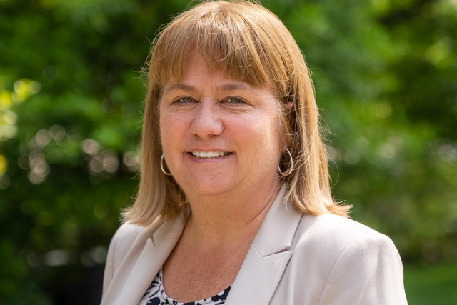 Sue Porterfield, a white woman with short light brown hair, in a beige jacket and floral shirt, standing outdoors in front of greenery.