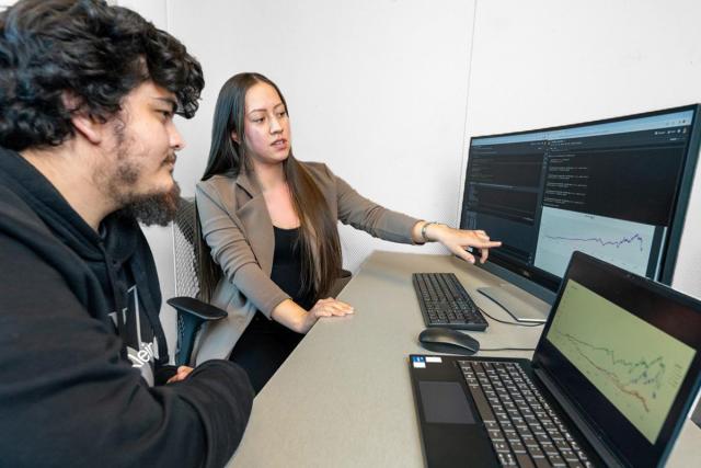 A young man and women sit before a computer at a desk. The woman is pointing to the screen.