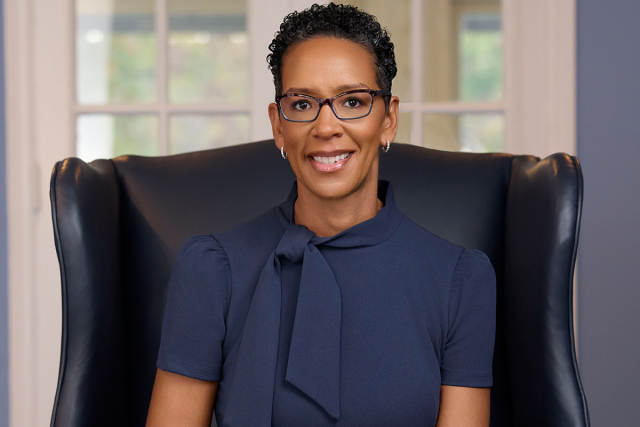 Darlene Repollet, with short black hair and glasses and wearing a dark blue suit, smiles as she sits in an upholstered chair.