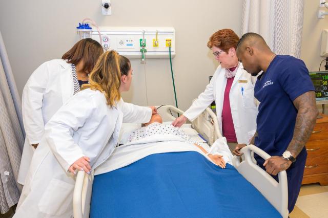 Three women and one man in nursing uniforms around a bed with a mannequin patient.