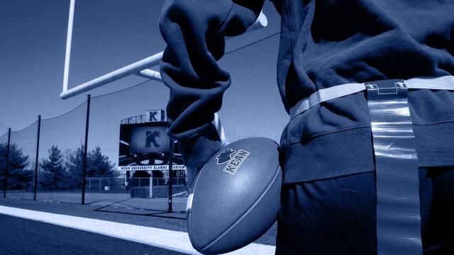 Upclose photo of a flag football player holding a Kean University football on a field; blue.