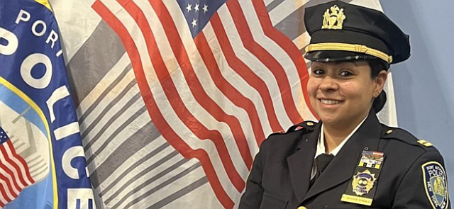 Smiling Hispanic woman in a black Port Authority uniform, including a hat, in front of a US flag